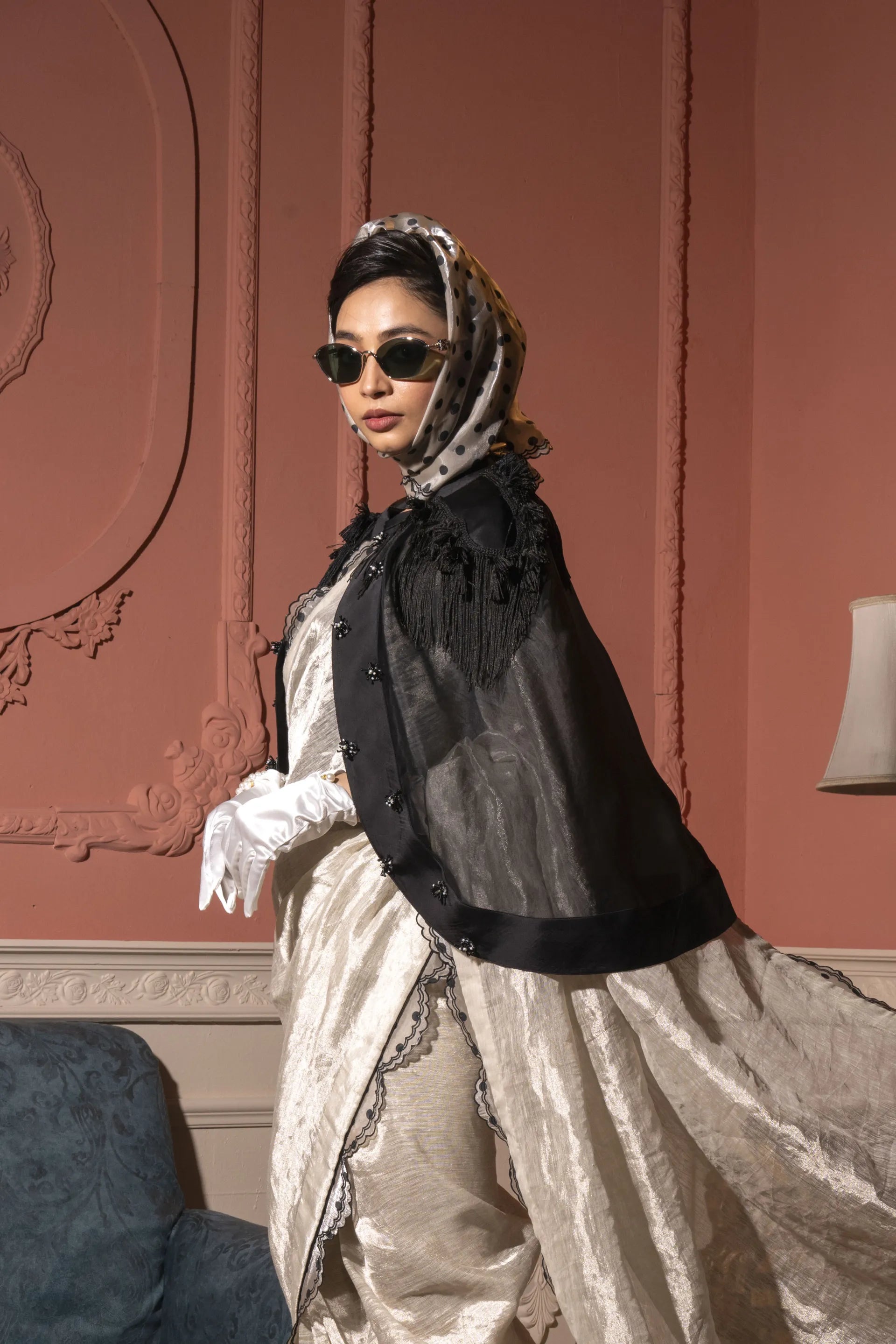 Person wearing a vintage outfit with a black coat and white dress against a decorative wall.